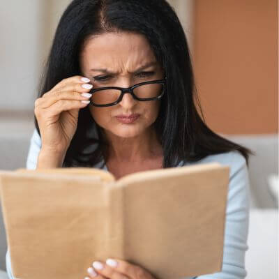 Middle-aged dark-haired woman pulling her glasses down to read a book more clearly
